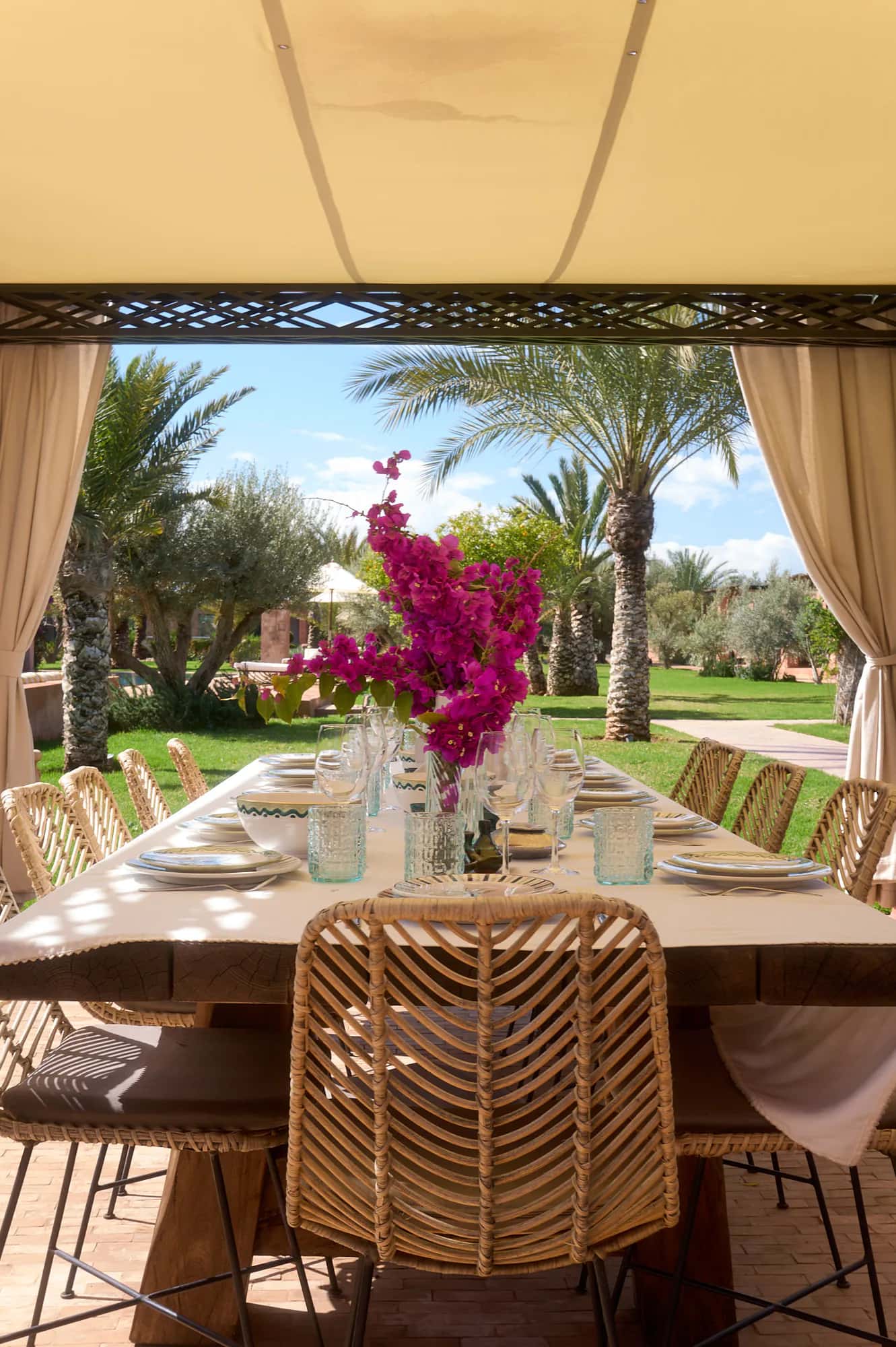 Large Villa Yemna outdoor dining table under a pergola, with pink bougainvillea, rattan chairs, and palm garden views.