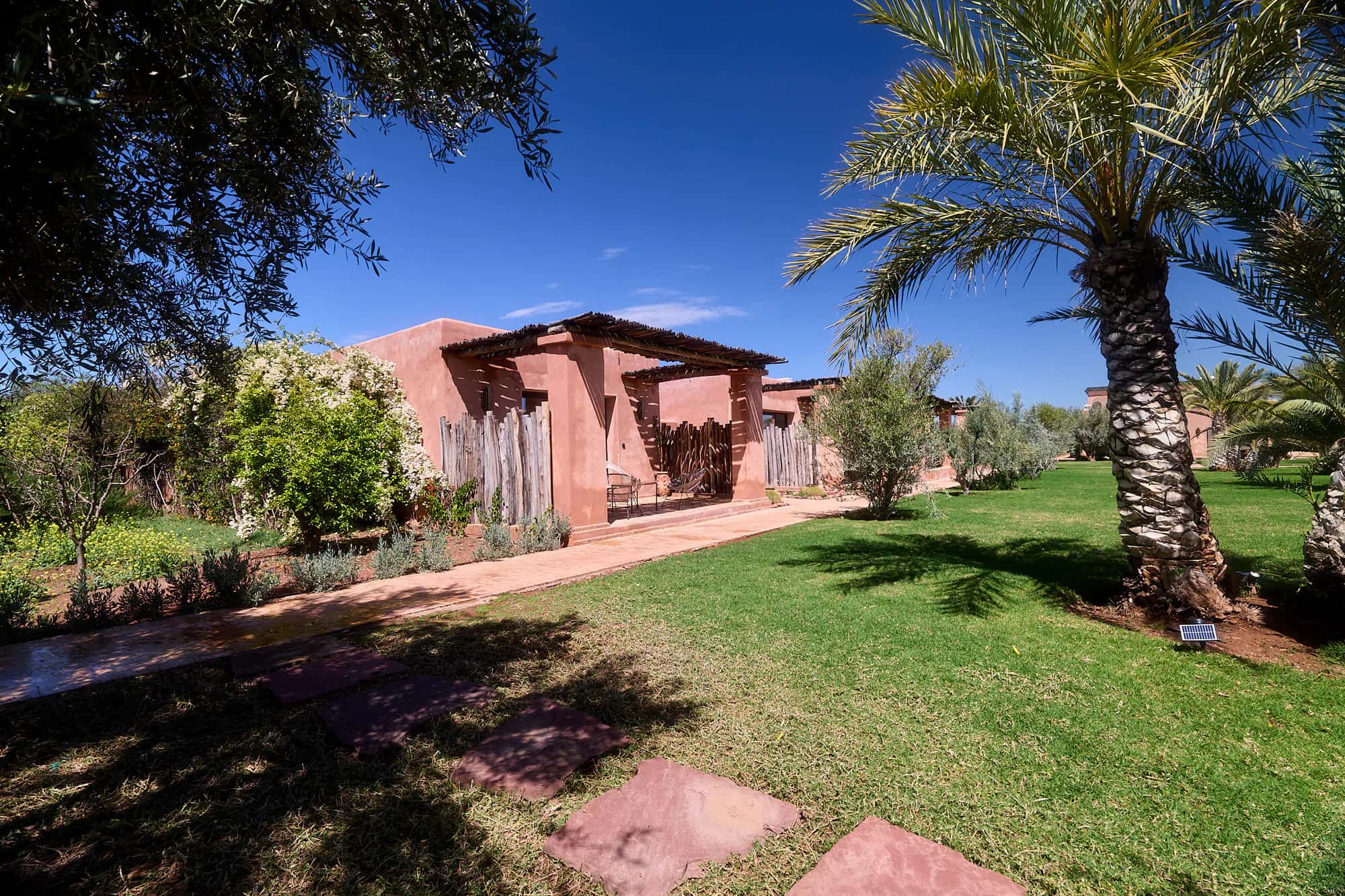 Villa Yemna garden path leading to a bungalow suite, with a palm tree, lawn, and Moroccan terracotta architecture.