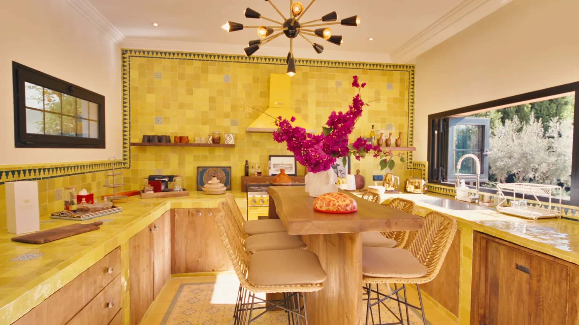 Villa Yemna riad kitchen in Marrakech with sunlit yellow zellige tiles, fuchsia bougainvillea, wooden counter and rattan stools.