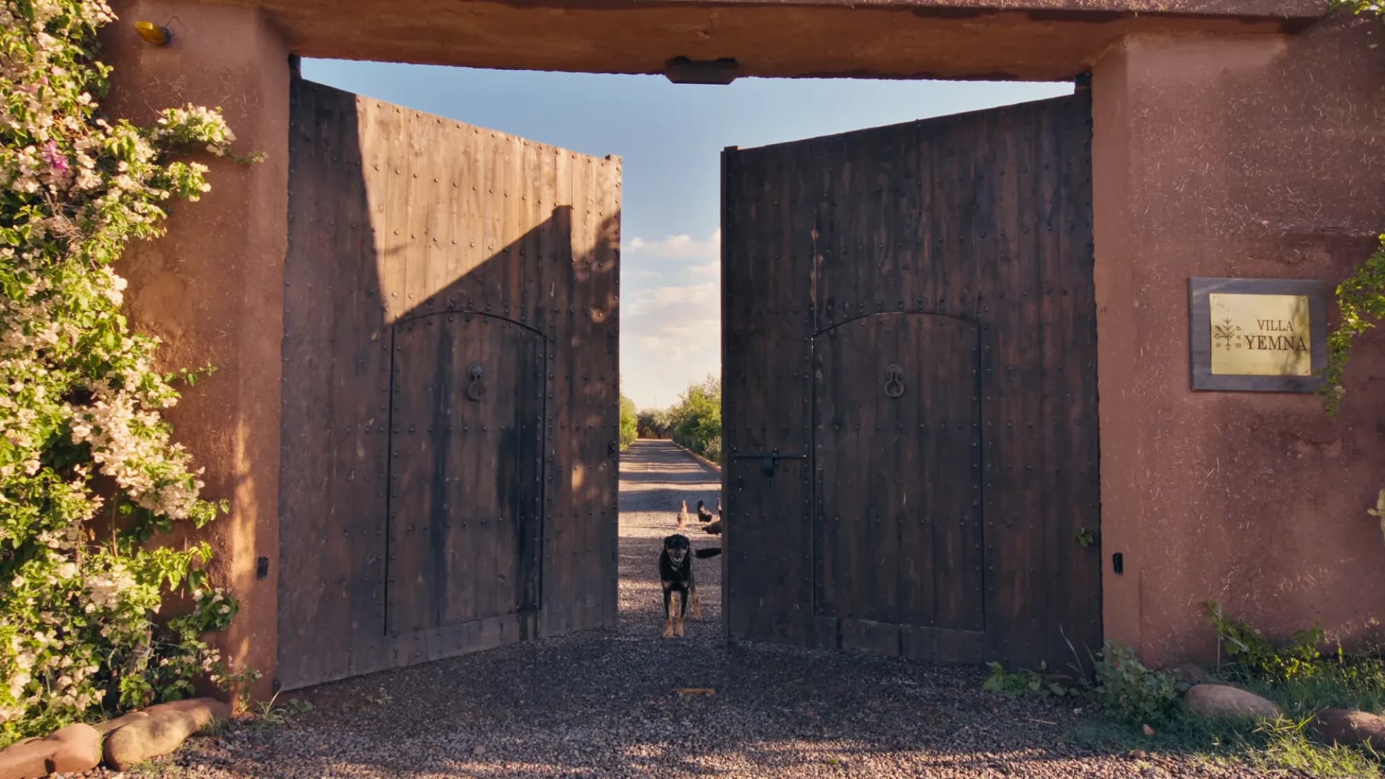 Wide open wooden entrance gate of Villa Yemna in Marrakech, with engraved signage and gravel driveway under golden light.