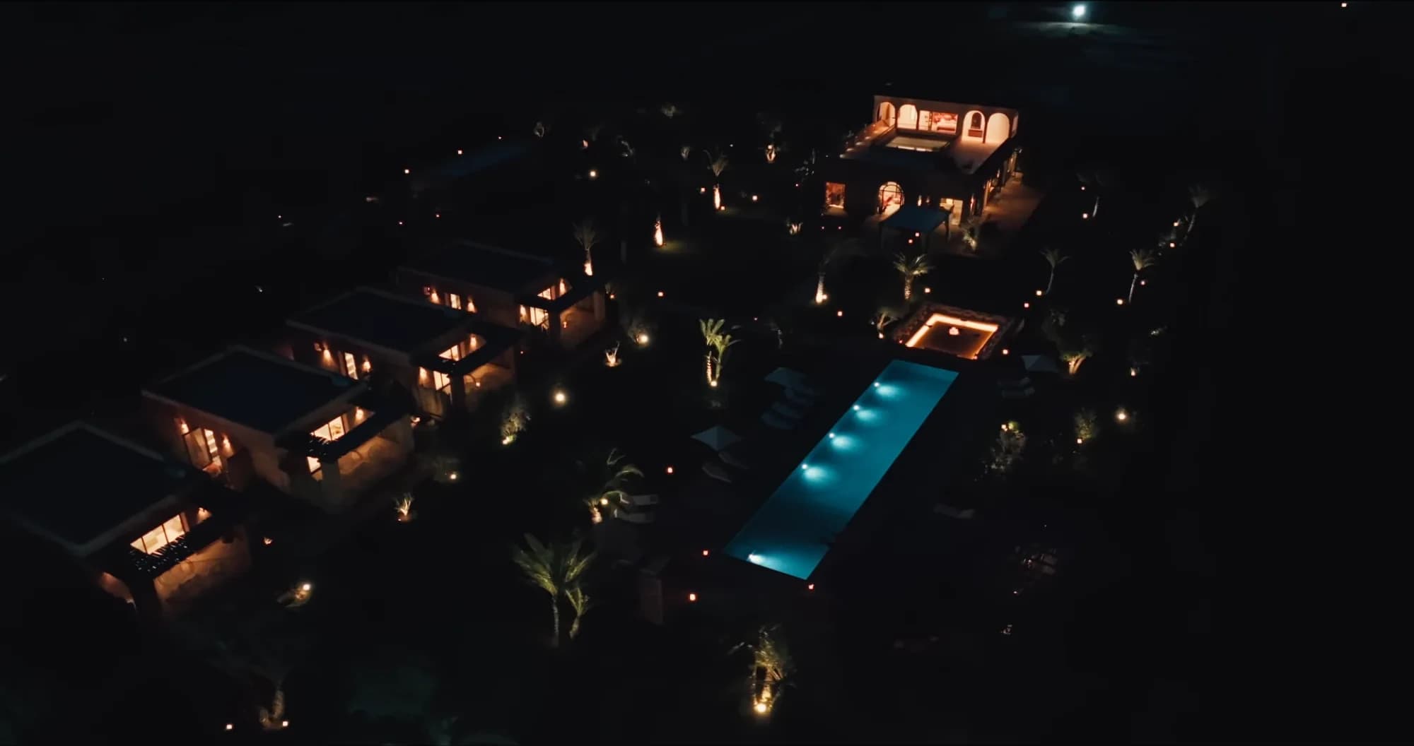 Aerial night view of the Villa Yemna estate in Marrakech illuminated: riad, bungalow suites, pool and gardens lit under the starry sky.