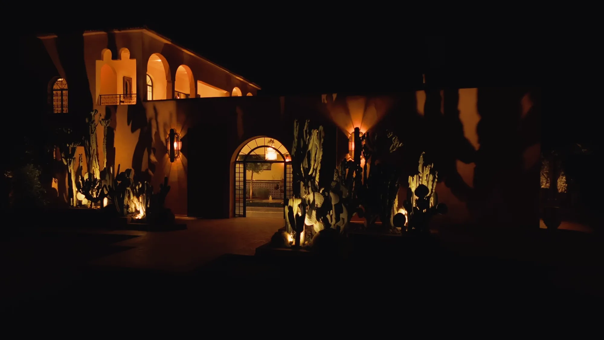 Villa Yemna façade at night in Marrakech with illuminated arched entrance, uplit cacti and bronze sconces.