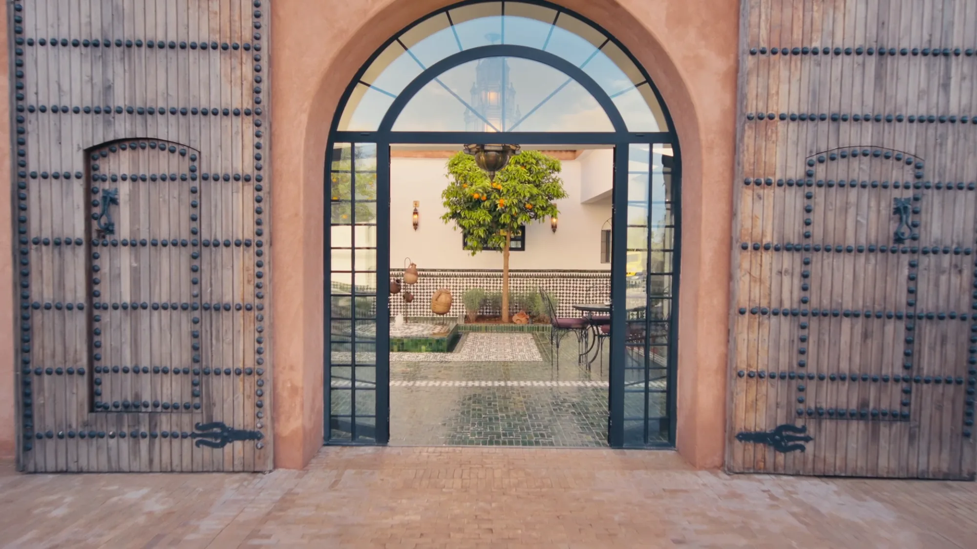 Studded wooden double doors opening onto the riad inner courtyard at Villa Yemna in Marrakech, with potted orange tree and green zellige tiles.