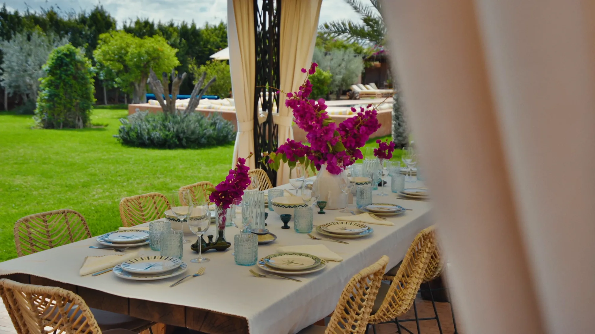 Table extérieure de Villa Yemna à Marrakech dressée pour un repas, avec bougainvilliers fuchsia, chaises en rotin et vue sur le jardin.