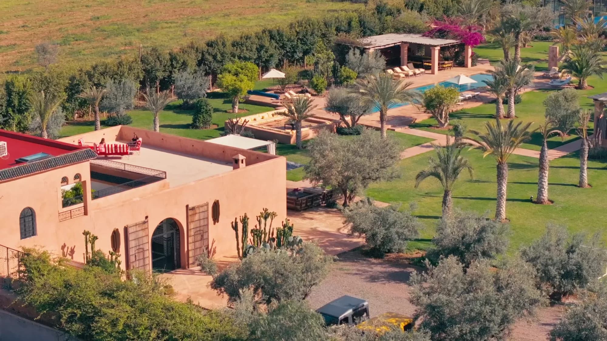 Vue aérienne du riad de Villa Yemna à Marrakech avec pergola adjacente, piscine, oliveraie et toit en terrasse au coucher du soleil.