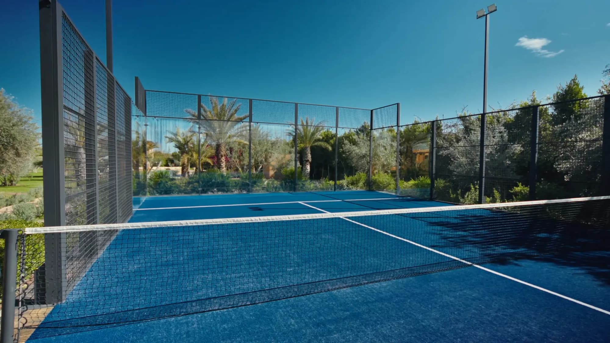 Terrain de padel privé de Villa Yemna à Marrakech vu depuis l'intérieur, avec surface bleue, filet et oliveraie environnante.