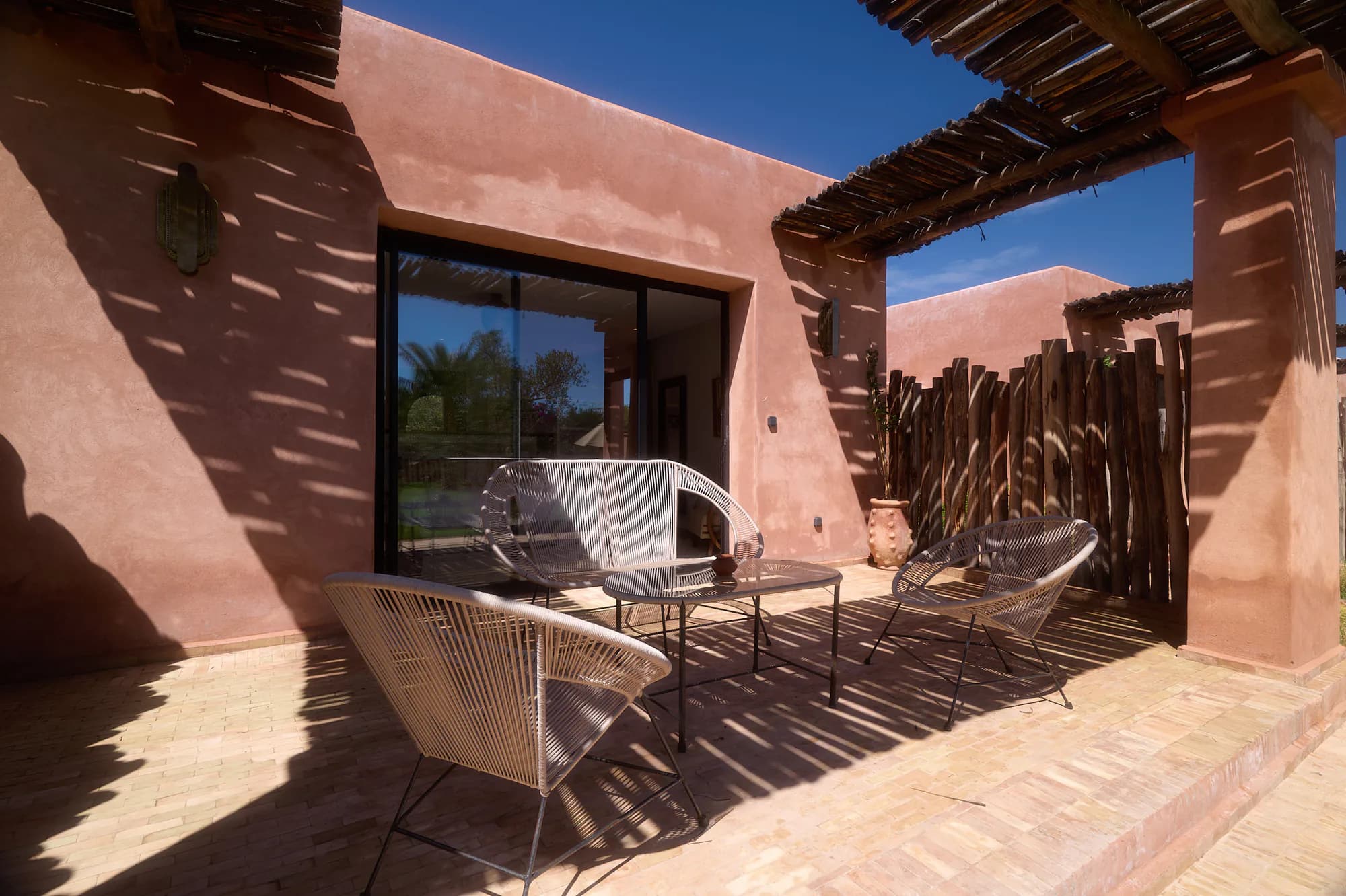 Terrasse privée d’une suite bungalow de Villa Yemna avec fauteuils tressés, table basse, pergola d’ombrage et murs terracotta.