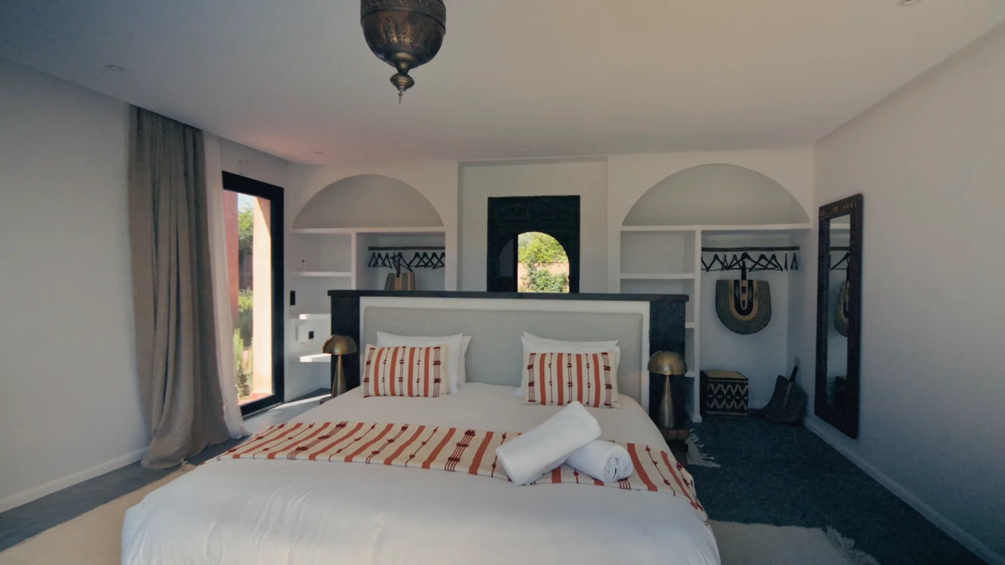 Chambre d’une suite de Villa Yemna à Marrakech avec lit king size en linge blanc, niches arquées, coussins rayés terracotta et suspension marocaine.