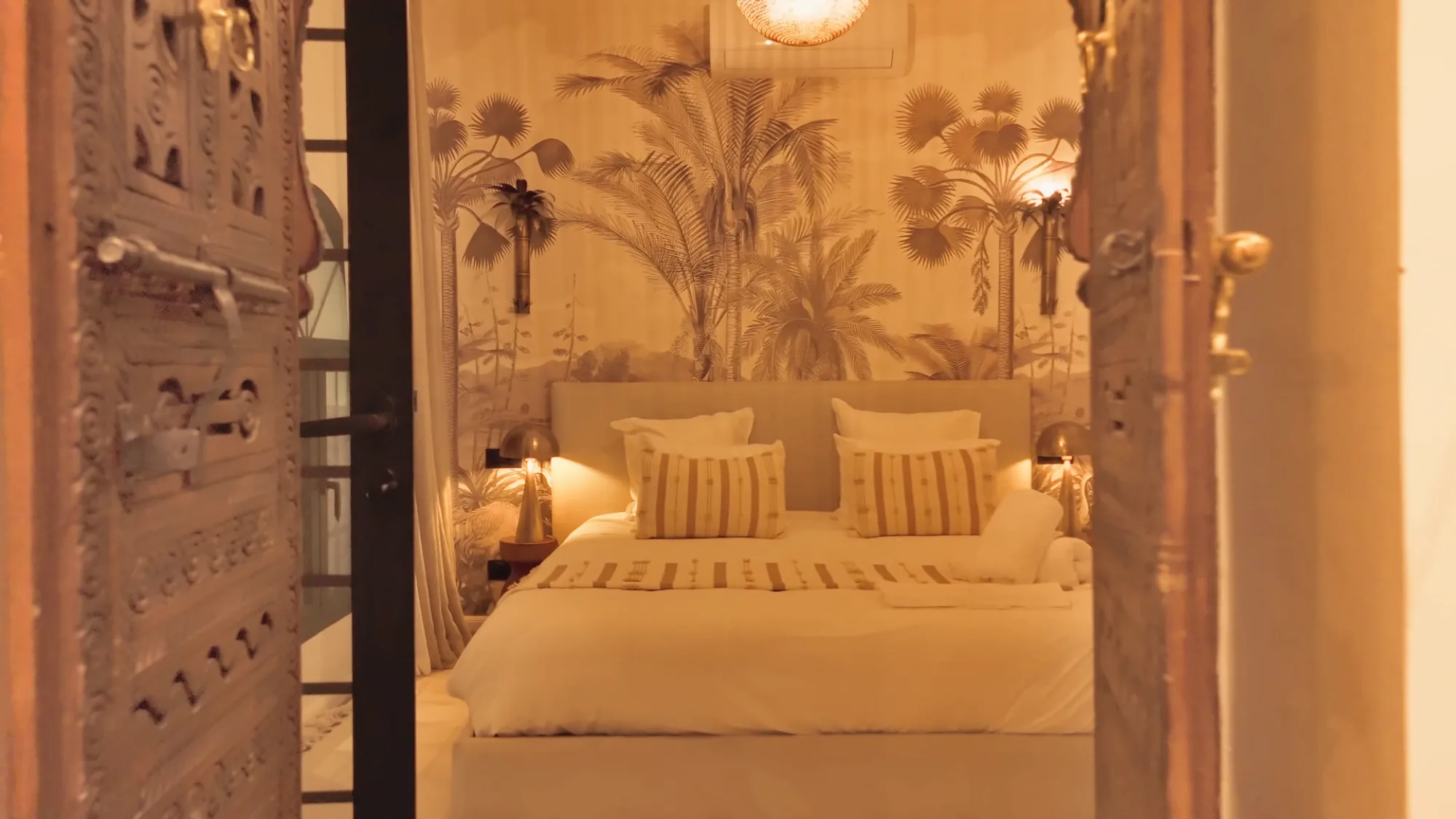 Villa Yemna suite bedroom in Marrakech with palm tree wall mural, white linen bed with striped pillows, viewed through carved Moroccan doors.