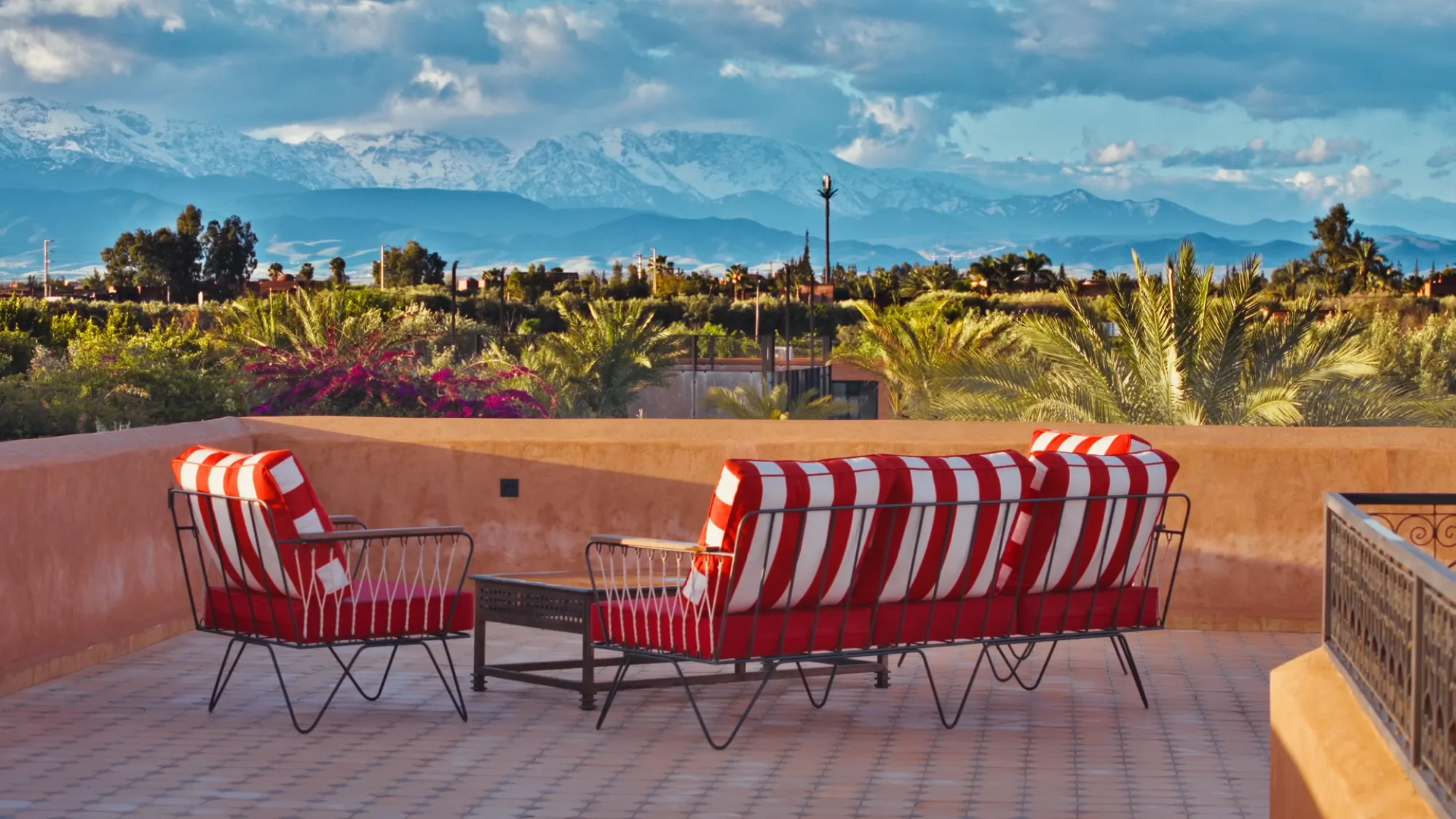 Villa Yemna rooftop terrace in Marrakech with red and white striped outdoor furniture, view of the snow-capped Atlas Mountains and palm trees.