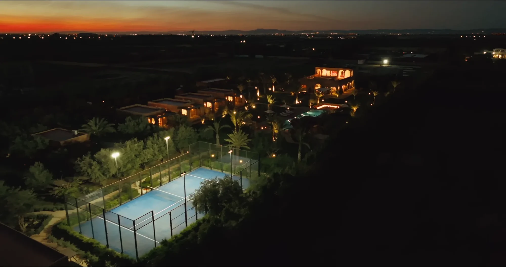 Vue drone au crépuscule de Villa Yemna à Marrakech avec terrain de padel éclairé, piscine et ligne d'horizon orangée sur la plaine.