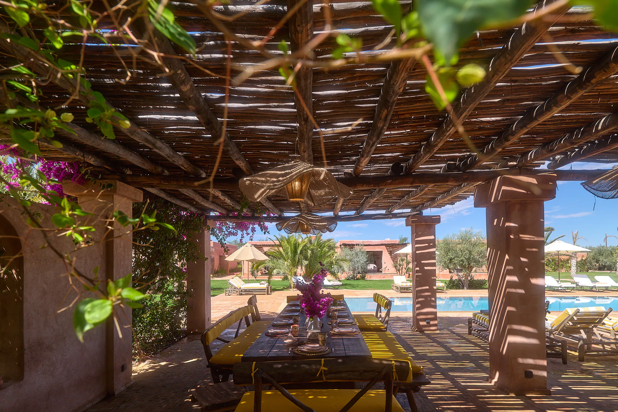 Espace repas ombragé sous pergola à Villa Yemna, avec table dressée, bougainvilliers et vue sur la piscine et les jardins.