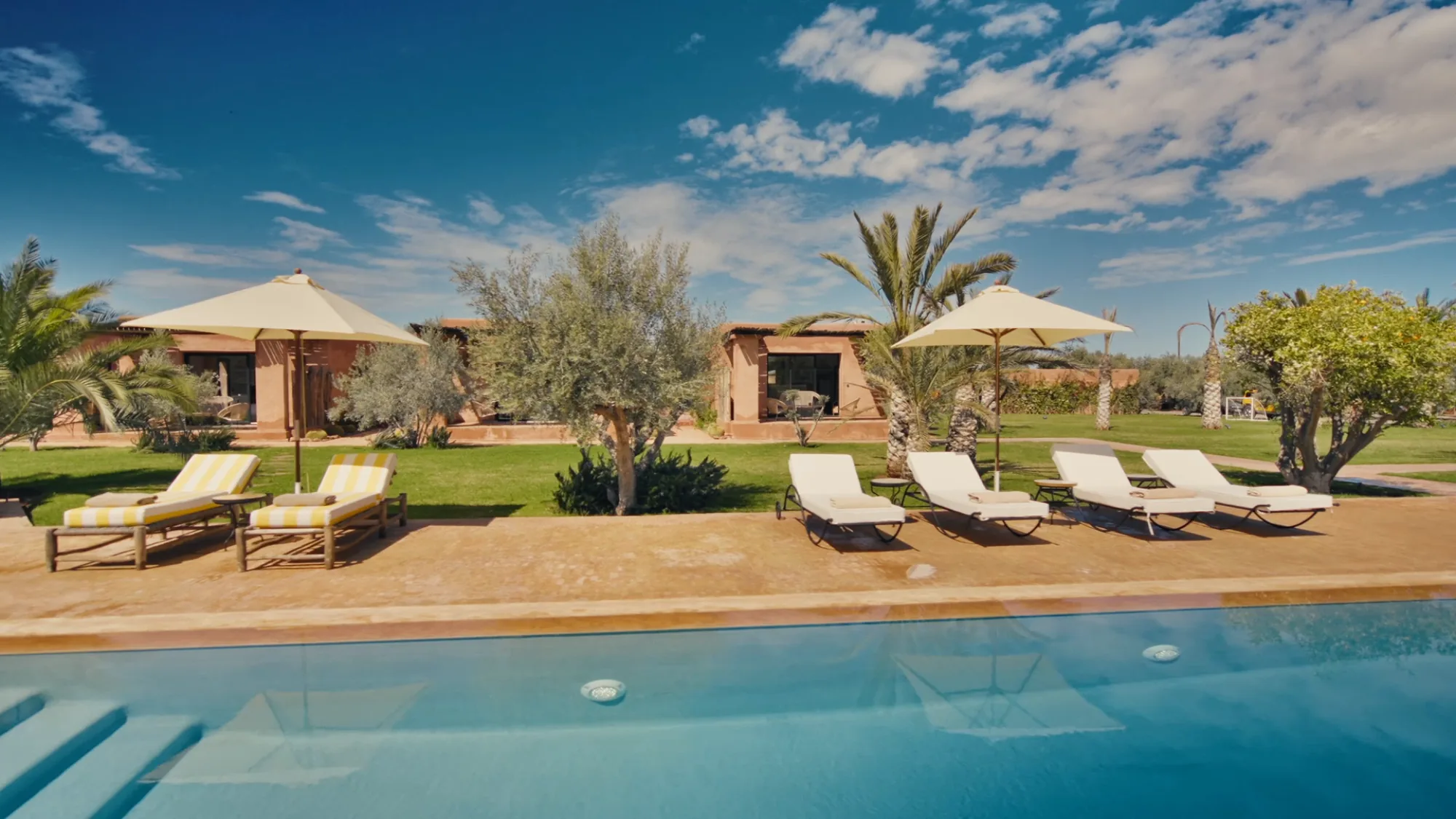 Deck de la piscine de Villa Yemna à Marrakech avec transats, parasols, oliviers et bungalow d'une suite en arrière-plan.