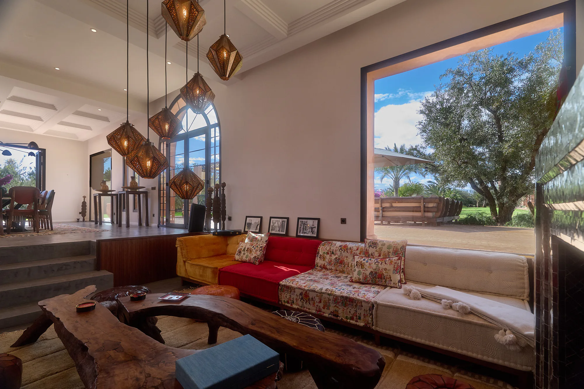 Salon du riad de Villa Yemna avec canapés à motifs, lanternes suspendues, cheminée en zellige et grande fenêtre ouverte sur le jardin.
