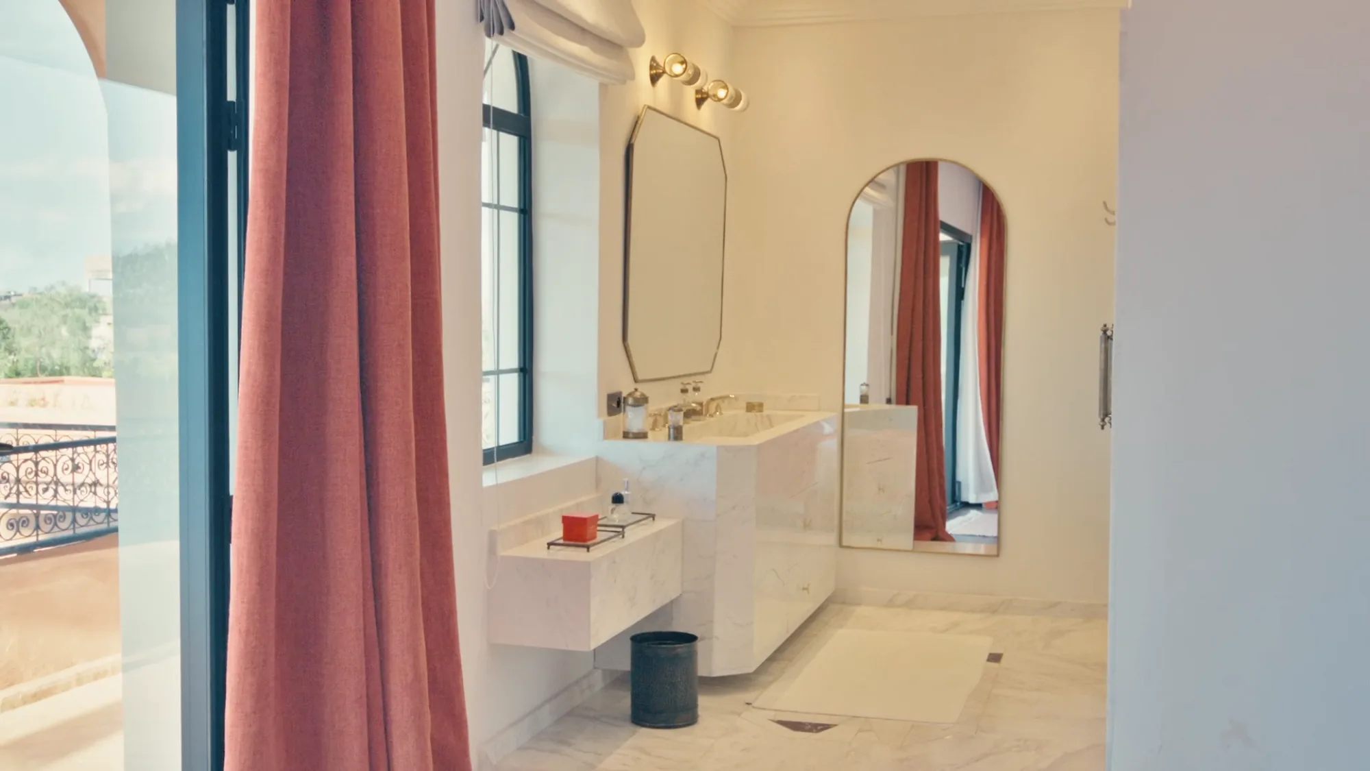 Salle de bain du riad de Villa Yemna à Marrakech avec double vasque en marbre, rideau terracotta, miroirs arqués et balcon en fer forgé.