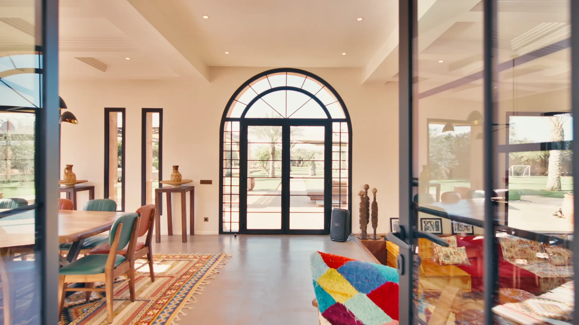 Salon du riad de Villa Yemna à Marrakech avec grande baie arquée, canapé multicolore patchwork, tapis berbère et sculptures africaines.
