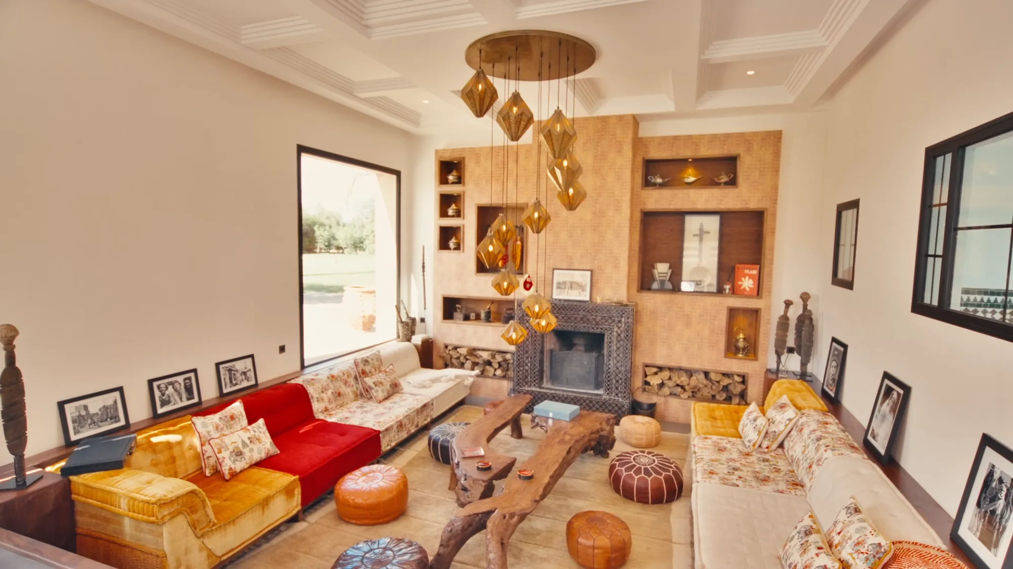 Salon du riad de Villa Yemna à Marrakech avec canapé rouge, cheminée en zellige, poufs en cuir et grappe de lampes suspendues en cuivre.