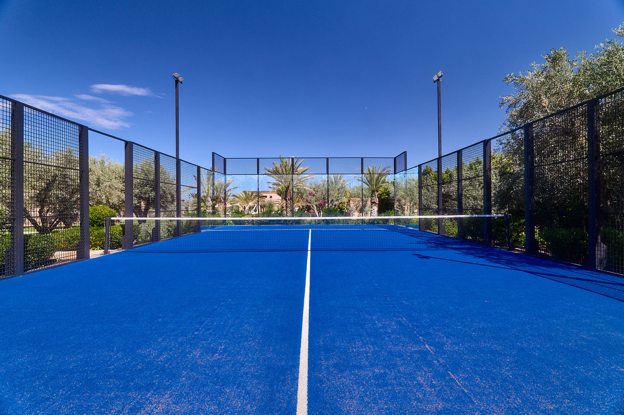 Terrain de padel bleu de Villa Yemna vu depuis la ligne centrale, entouré de vitres, d'oliviers et de palmiers.