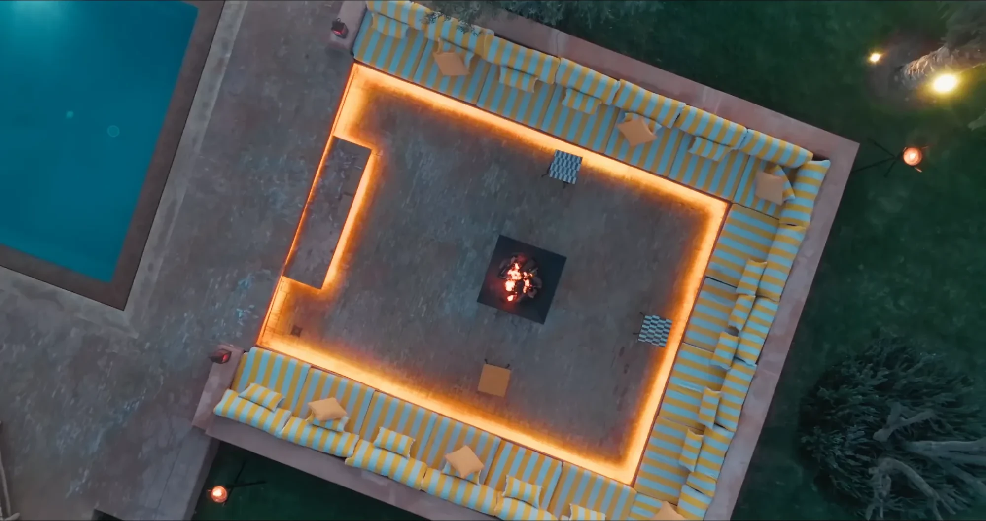 Vue plongeante drone de l'espace brasero de Villa Yemna à Marrakech au crépuscule, avec lounge en L, coussins rayés bleus et feu central allumé.