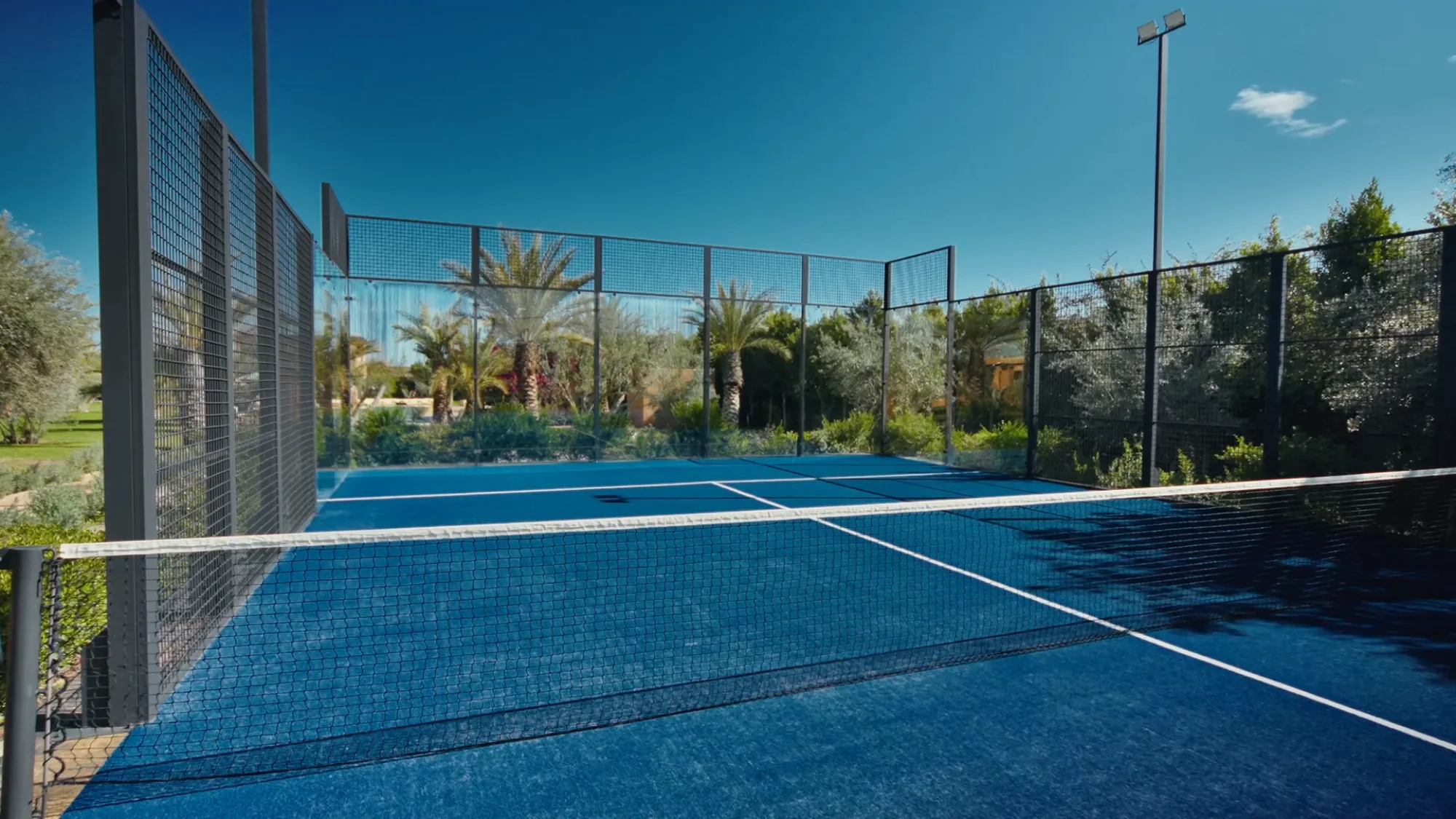 Terrain de padel privé de Villa Yemna à Marrakech vu depuis l'intérieur, avec surface bleue, filet et oliveraie environnante.