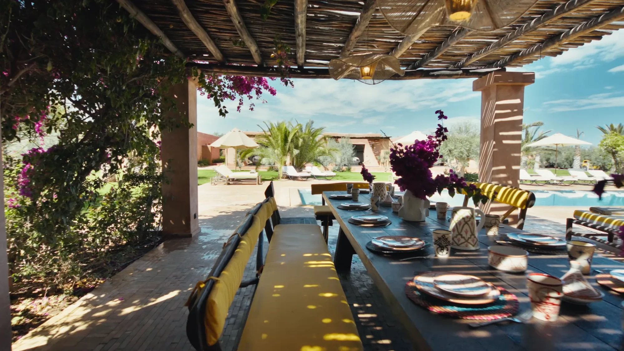 Table de repas sous pergola au bord de la piscine de Villa Yemna à Marrakech, avec coussins rayés, bougainvilliers et vue sur les jardins.