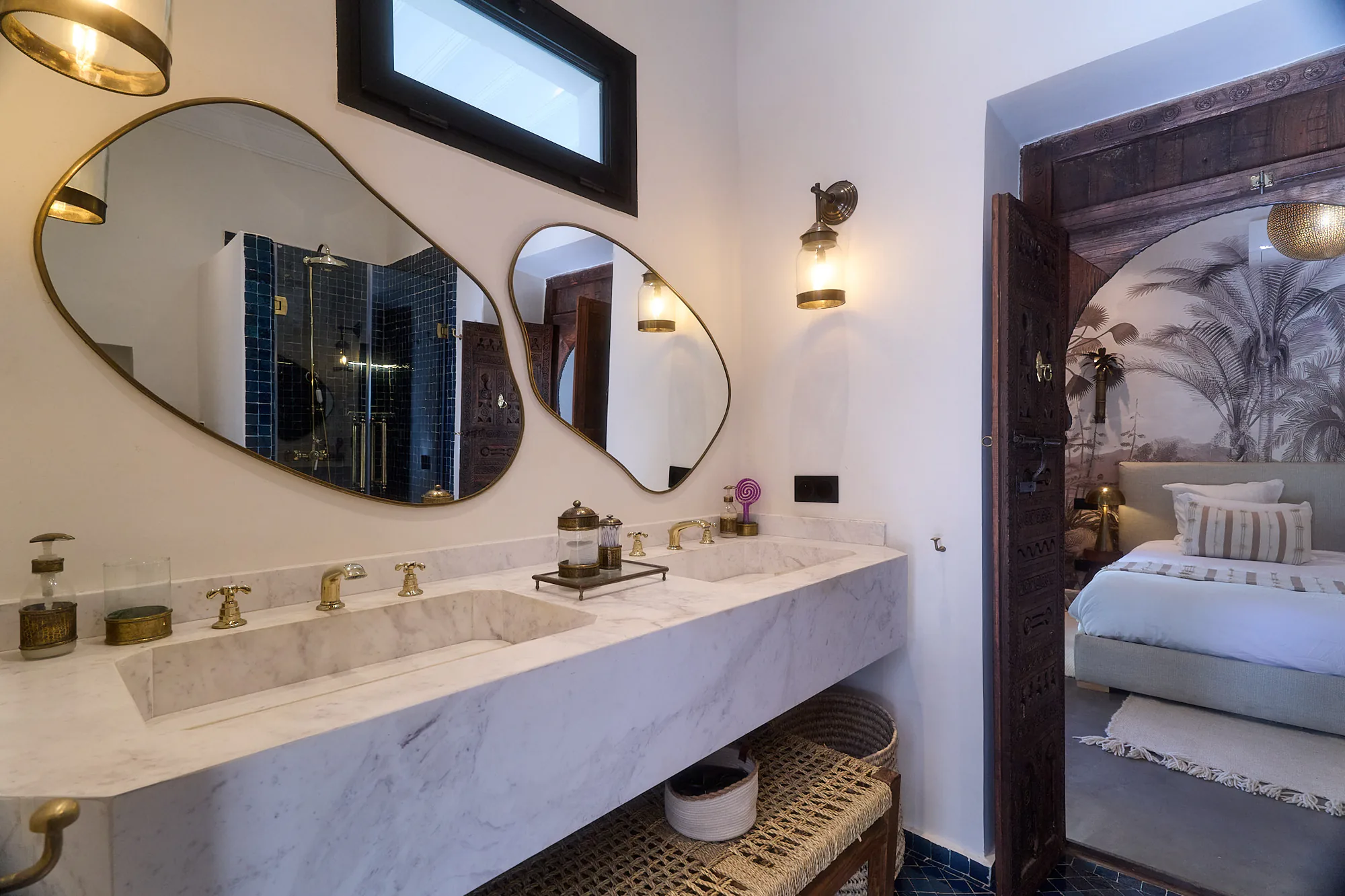 Salle de bain de la Suite Imsouane à Villa Yemna avec doubles miroirs organiques, vasque en marbre et vue vers la chambre.