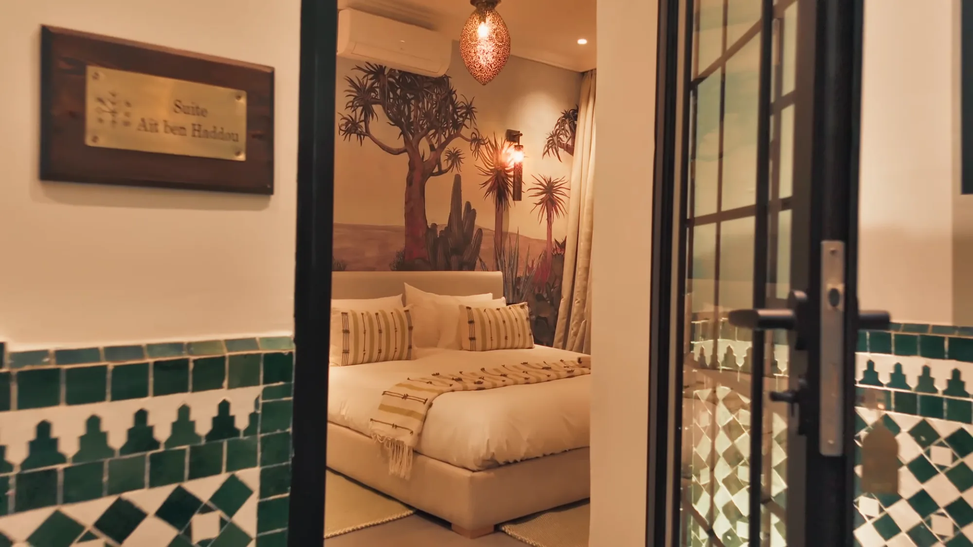 Entrée de la Suite Aït ben Haddou à Villa Yemna à Marrakech, avec zelliges verts, fresque de cactus, lit en linge crème et suspension marocaine.