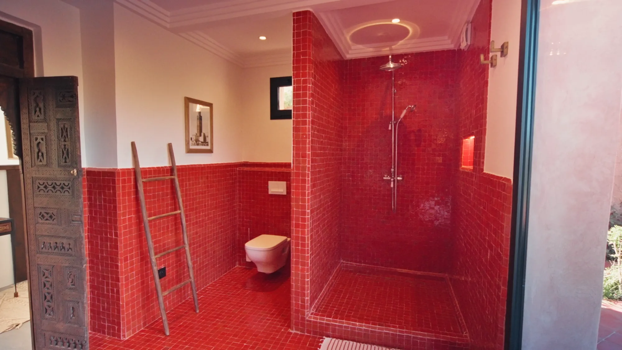 Salle de bain d'une suite de Villa Yemna à Marrakech avec zelliges rouges, douche à l'italienne, porte sculptée marocaine et échelle en bois.