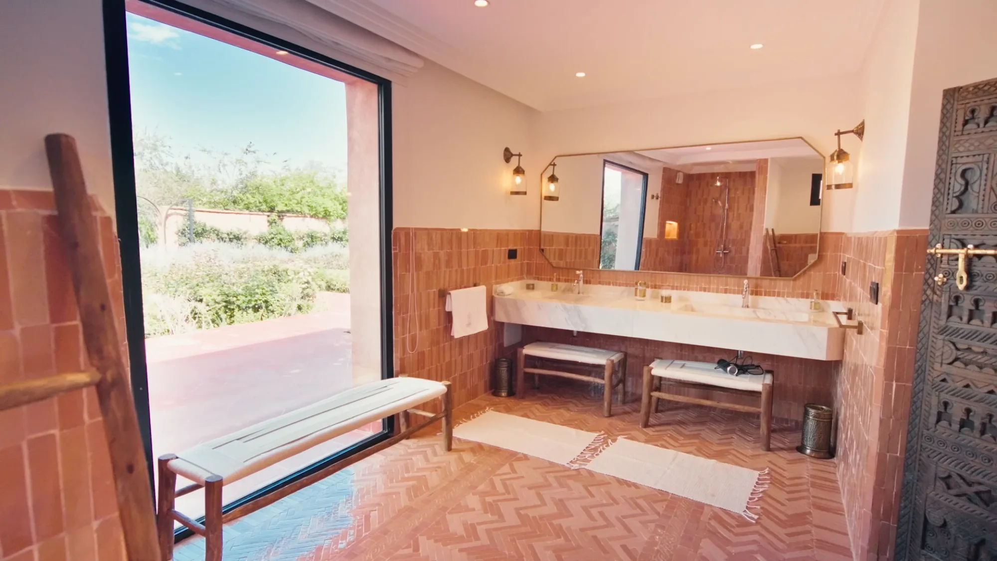 Salle de bain d'une suite de Villa Yemna à Marrakech avec carreaux terracotta, sol chevron, double vasque marbre et grande ouverture sur le jardin.