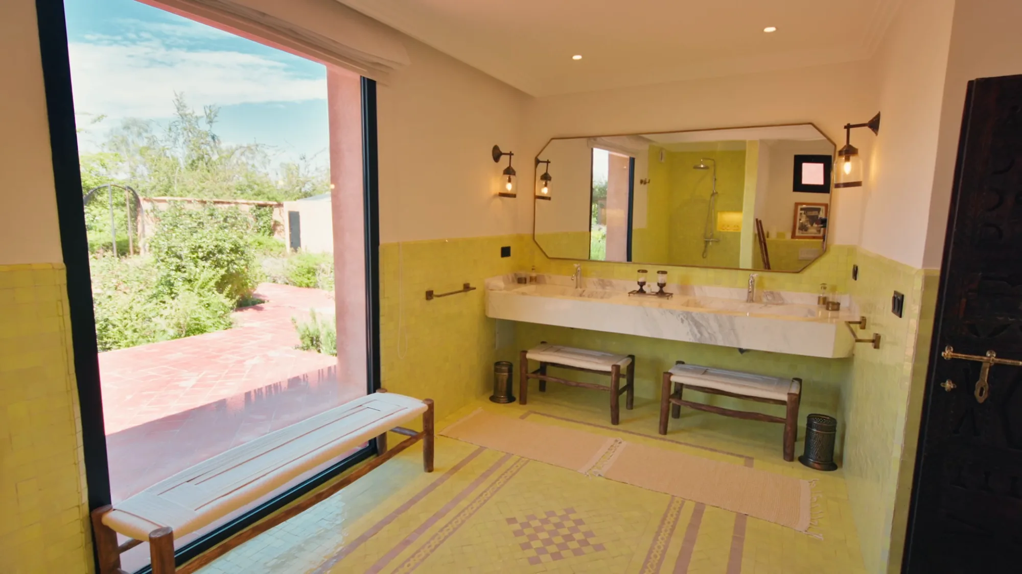 Salle de bain d'une suite de Villa Yemna à Marrakech avec zelliges jaunes, double vasque en marbre, miroirs arqués et vue sur le jardin.
