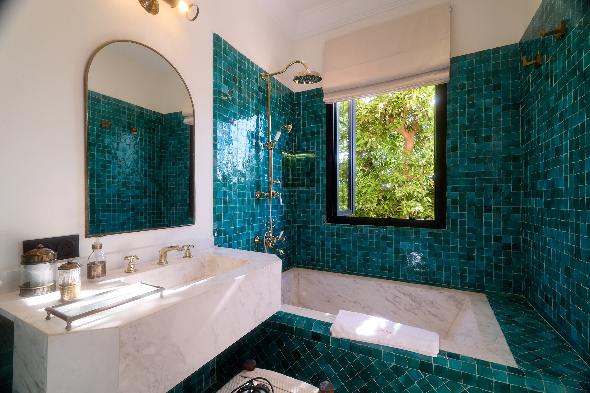Salle de bain de la Chambre Taghazout à Villa Yemna, avec zelliges bleu-vert, baignoire en marbre, douche laiton et fenêtre sur la verdure.